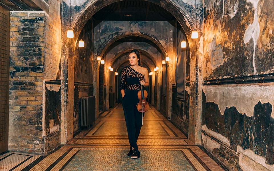Nicola Benedetti holding violin and looking towards the side, standing in a corridor