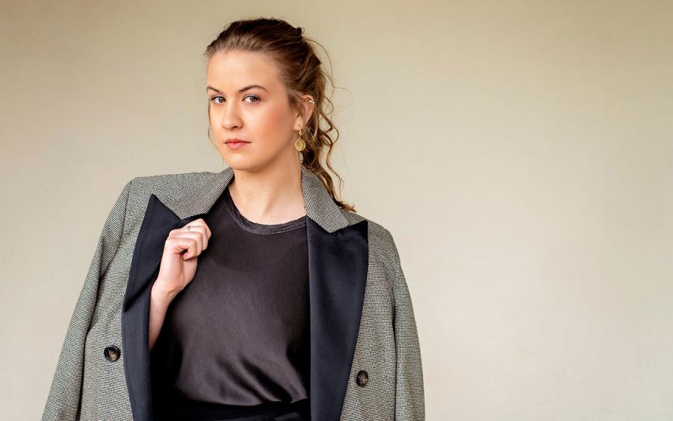 Charlotte Corderoy against a light coloured wall. She has her hair tied back, gold earrings, and is looking directly to the camera. She is wearing a dark top with a jacket over her shoulders.