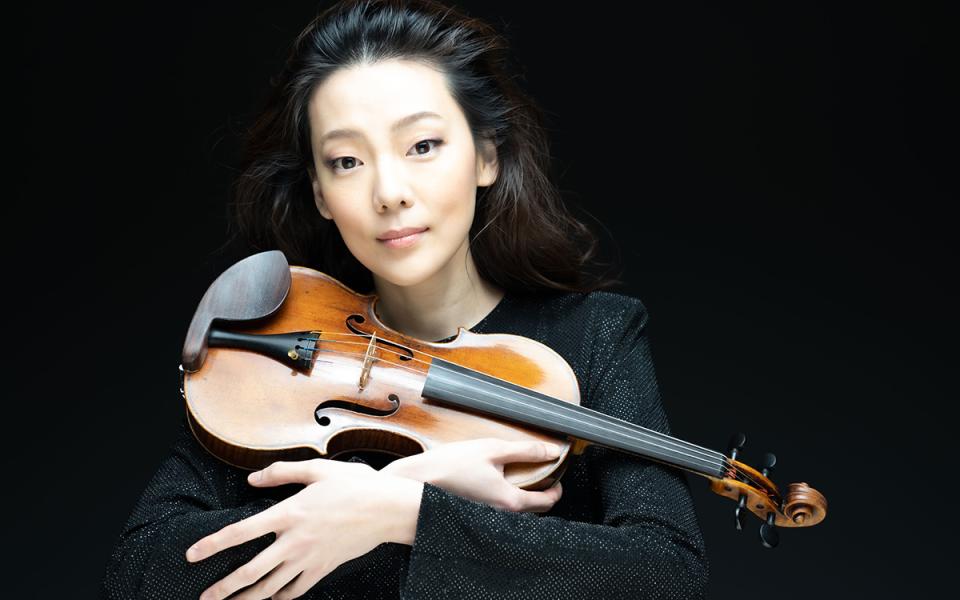 Clara-Jumi Kang against a black background, looking into the camera. She is resting her violin across her body