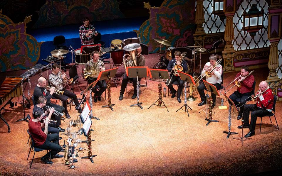 Brass players sat in a semi circle wearing casual clothes and Christmas jumpers. They are sat amongst a colourful pantomime set.
