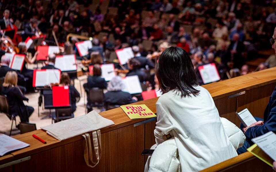 Audience member leaning over the choir stalls at Royal Festival, watching the Philharmonia Orchestra. The camera focuses on a Philharmonia concert programme